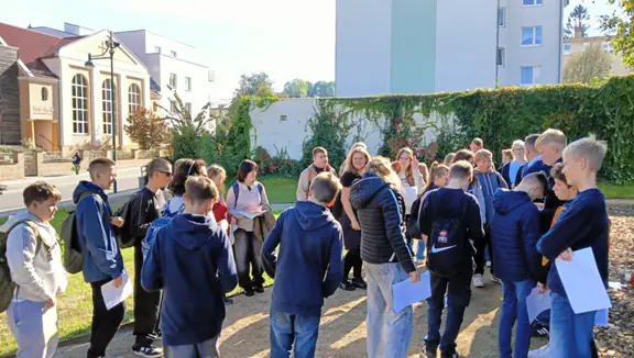 Grupa uczniów jest na spacerze w mieście i w swobodnej grupie słuchają nauczyciela.