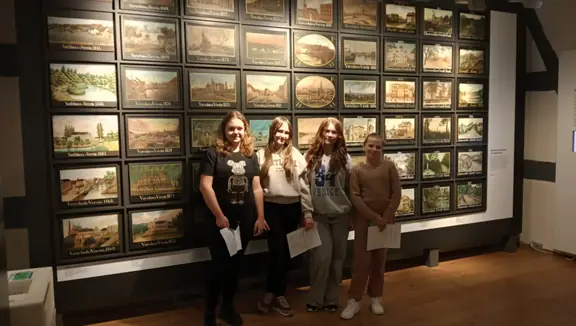 Cztery uczennice ze Stargardu pozują w muzeum przy ścianie ozdobionej starymi pejzażami miasta. 