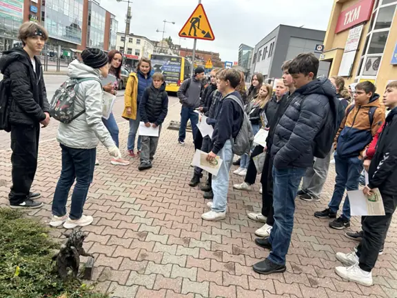 Uczniowie stoją na ulicy Wyszyńskiego, przy gryfiku i słuchają opisu.