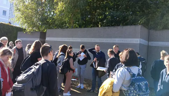 Grupa uczniów stoi przy budynku, który symbolizuje miejsce dawnej synagogi. Uczniowie słuchają przewodnika.