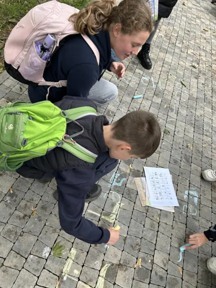 dwoje uczniów patrzy na kartkę i pisze według niej swoje imię na chodniku