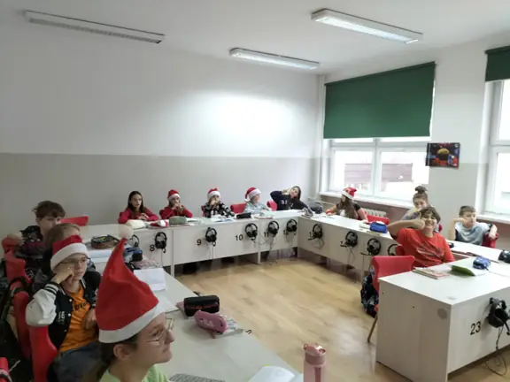 Uczniowie z kl. IVa siedzą przy stolikach w mikołajkowych czapeczkach.