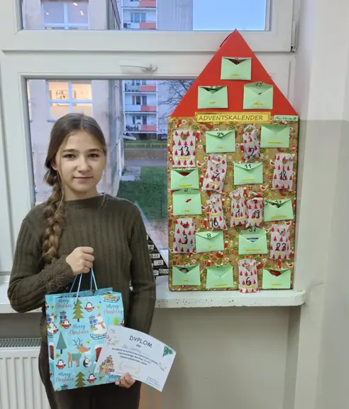 Uczennica pozuje do zdjęcia z dyplomem i nagrodą, a w tle stoi kalendarz przez nią wykonany