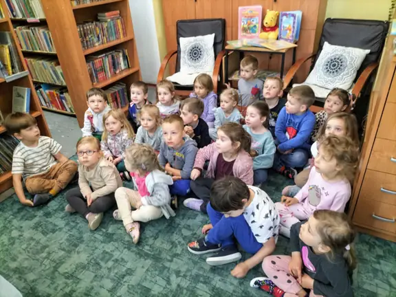 Przedszkolaki siedzą na dywanie w bibliotece i oglądają film - Jak powstaje książka.
