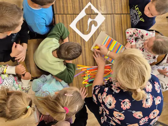 Pani bibliotekarka pokazuje dzieciom sposób przyklejania kolorowych pasków papieru.