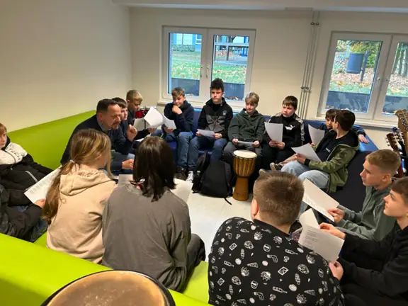 Grupa uczniów siedzi w kręgu i uczy się tekstu piosenki.