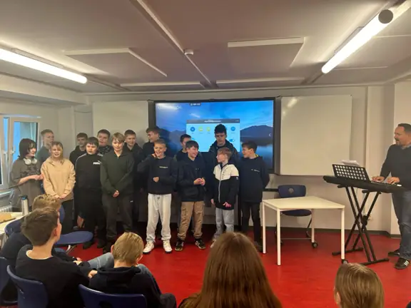 Grupka uczniów śpiewa piosenkę, stojąc przed wszystkimi. Obok nauczyciel gra na keybordzie.