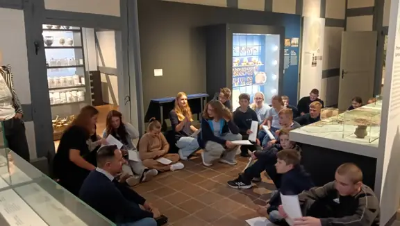 Uczniowie siedzą na podłodze w muzeum i słuchają nauczyciela. Dookoła widać niektóre eksponaty muzealne.